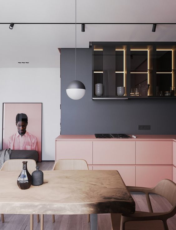 a chic grey kitchen with lower pink cabinets and upper black ones with built-in lights, a stained table and a pink artwork