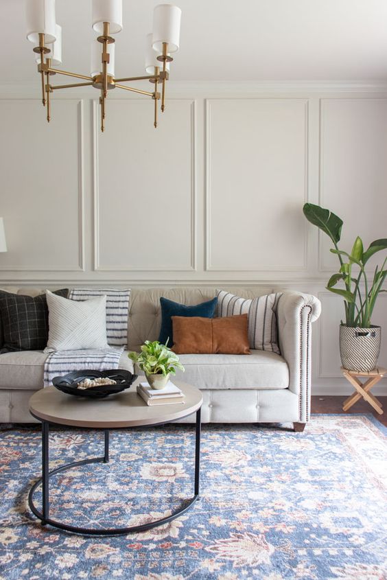 a chic living room with molding on the walls, a refined neutral sofa, a round table, a brass chandelier, mismatching pillows and a plant