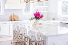 a contemporary glam kitchen with white cabinetry, white marble tiles, glam brass chandeliers and acryl and brass chairs