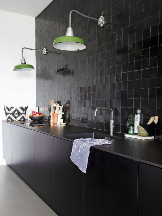 a cool black kitchen with sleek lower cabinets, tiles all over the wlal, green lamps and stone countertops