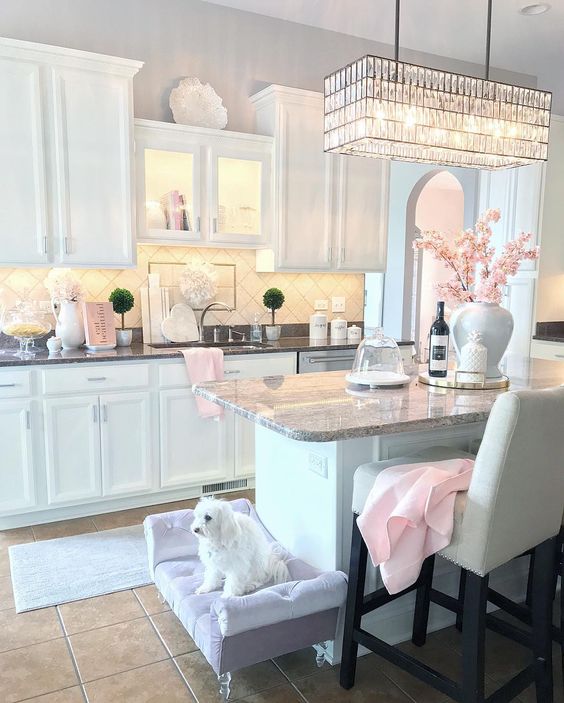 a cute glam kitchen with white cabinetry, stone countertops, a crystal chandelier, a lavender dog bed and touches of light pink