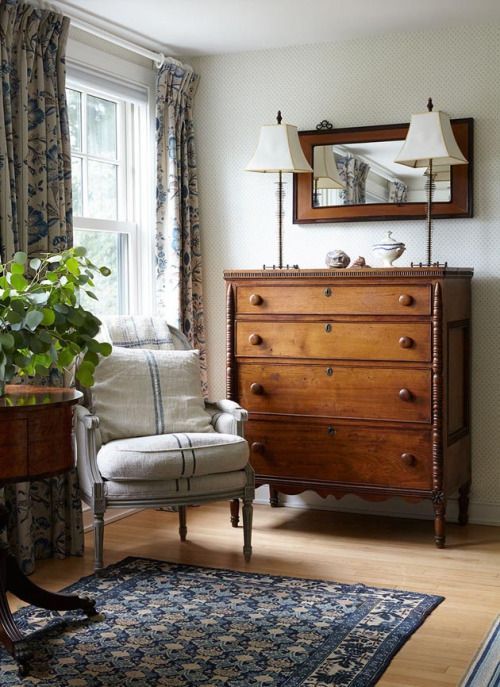 a farmhouse living room with a stained dresser, a whitewashed antique chair with pillows, printed textiles
