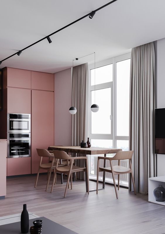 a laconic kitchen with grey walls and pink sleek cabinets, a stained table and chairs, black and white pendant lamps