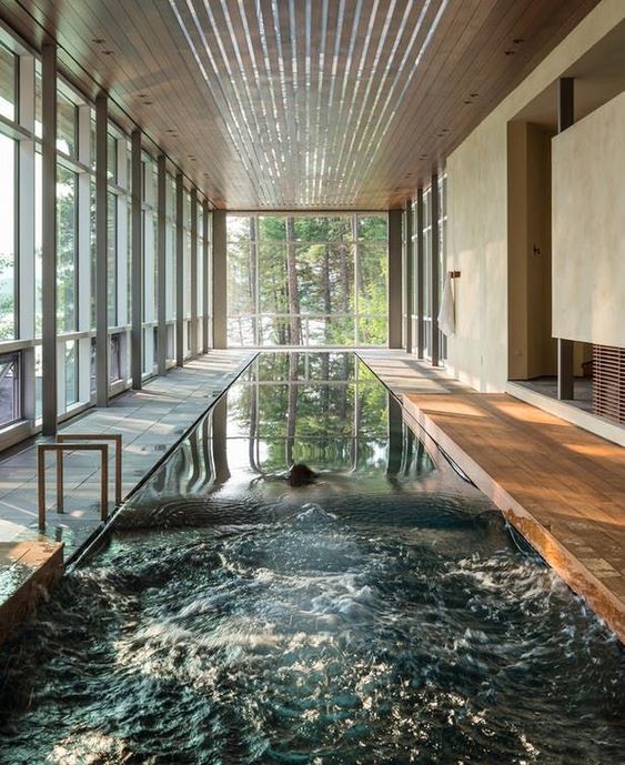 a large contemporary pool house with glazed walls and a wood slat roof, a large and long pool and a wooden deck plus forest views