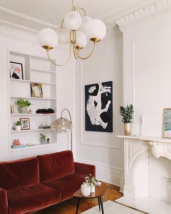 a lovely living room with molding on the walls, niche shelves, a burgundy velvet sofa, a non-working fireplace and cool artworks