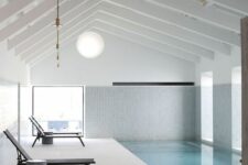 a minimalist pool house clad with neutral stone tiles and white ones, with a large pool and several loungers with side table