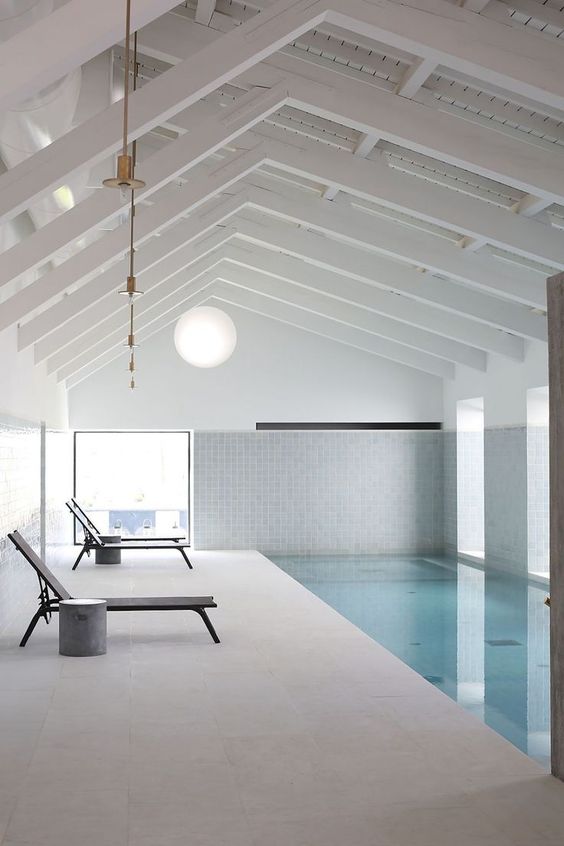 a minimalist pool house clad with neutral stone tiles and white ones, with a large pool and several loungers with side table