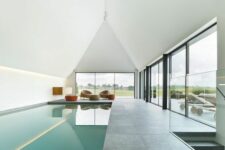a minimalist pool house with an attic roof and glazed walls, a large pool and a stone tile deck plus a sitting zone with rust stools and beige chairs
