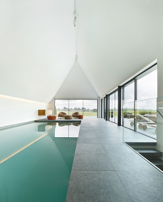 a minimalist pool house with an attic roof and glazed walls, a large pool and a stone tile deck plus a sitting zone with rust stools and beige chairs