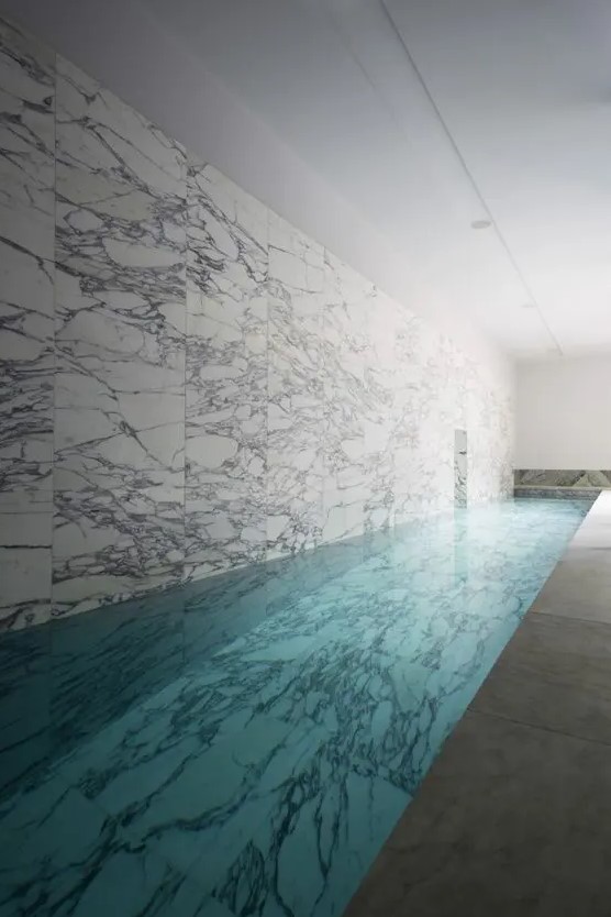 a minimalist yet luxurious indoor pool space clad with white marble, with stone tiles, long and quite a narrow pool