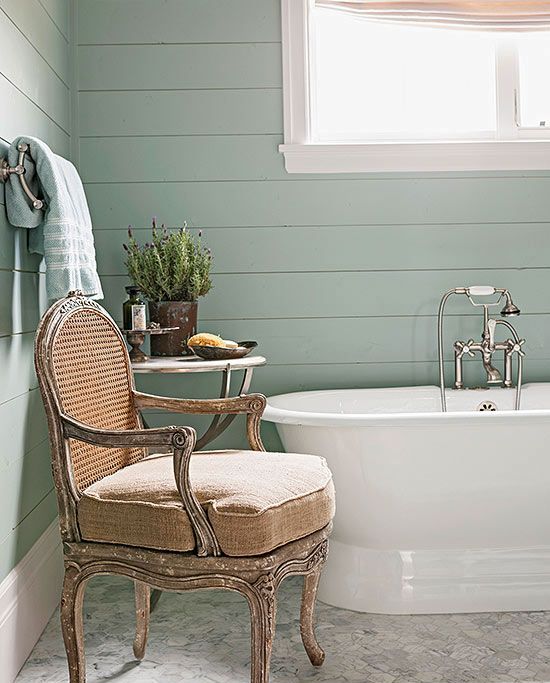 a mint green cottage bathroom with a vintage bathtub, a neutral antique chair, a side table with decor