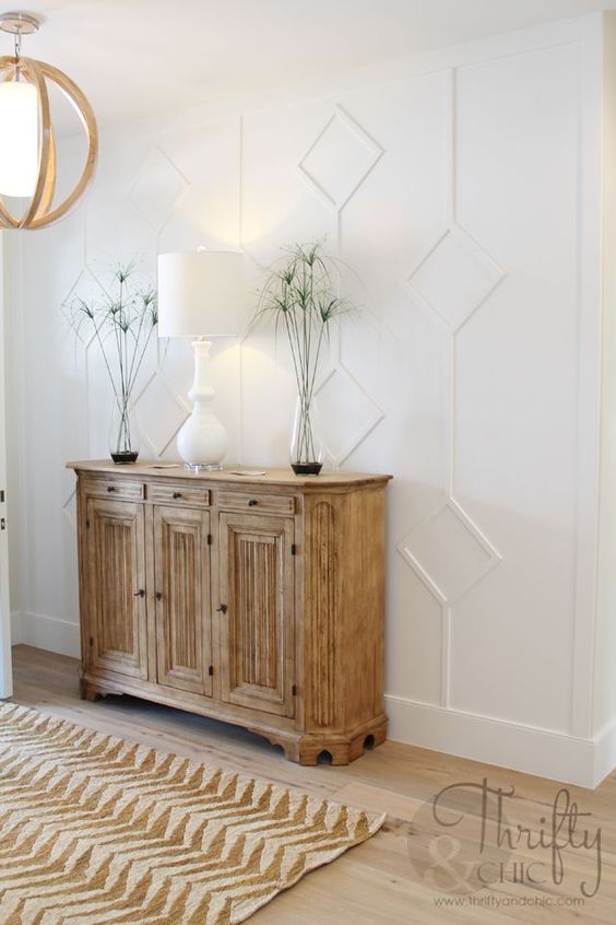 a modern farmhouse entryway with a white molded wall, a light-stained console, some greenery and a chic white lamp