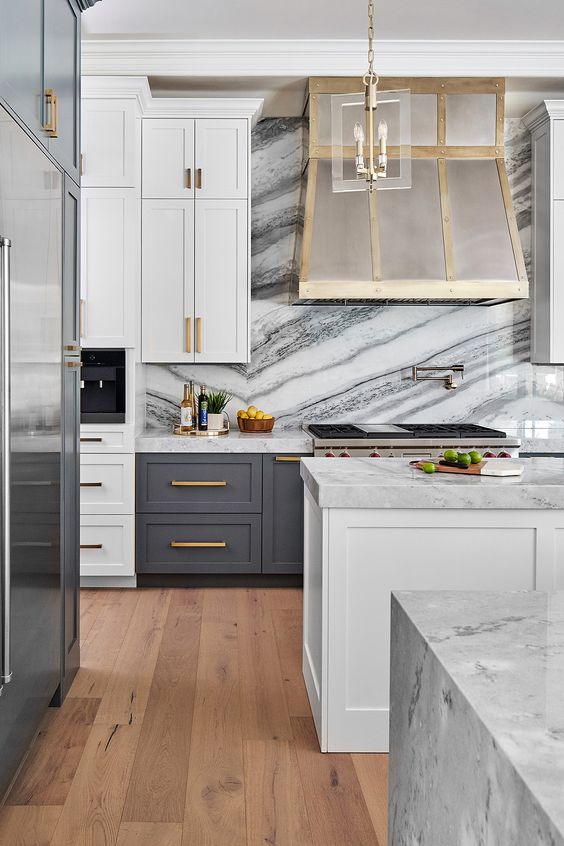 a modern glam kitchen with white and graphite grey cabinetry, a white stone backsplash and countertops, a glam hood and gold handles