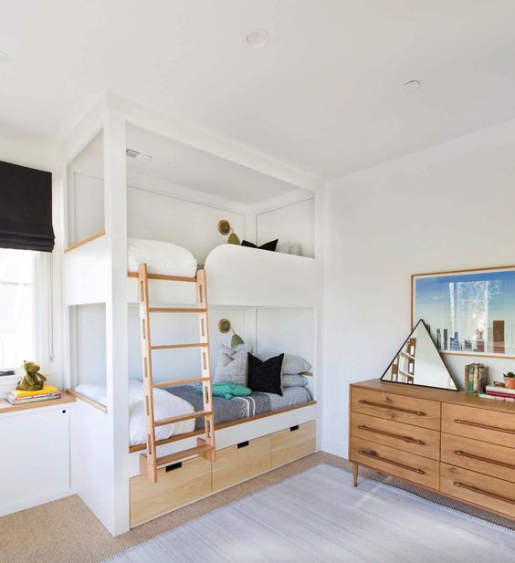 a modern kids' room with built-in bunk beds, with neutral bedding, a stained dresser, a triangle mirror, shelves and toys here and there