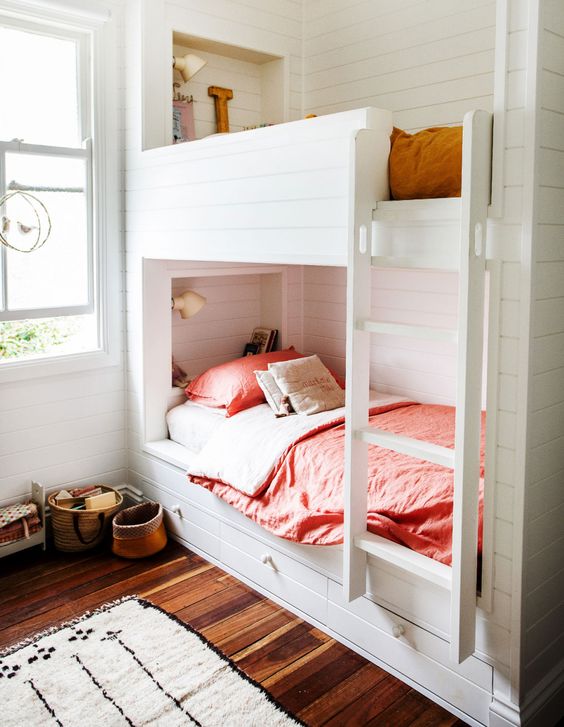 a modern neutral kids' room with white shiplap walls, built-in bunk beds with pink and mustard bedding, baskets and white sconces