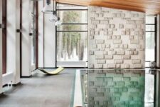 a modern pool house clad with stone tiles, with a stone accent wall and a stained sloped ceiling, a large pool and a series of pendant lamps