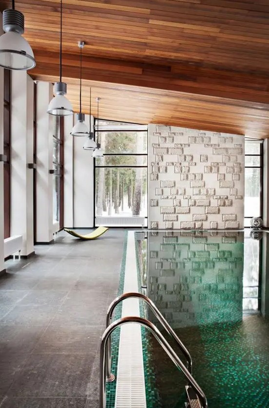 a modern pool house clad with stone tiles, with a stone accent wall and a stained sloped ceiling, a large pool and a series of pendant lamps