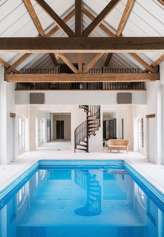 a modern pool house in white, with wooden beams, a large pool and a staircase plus a woven sofa to enjoy swimming anytime
