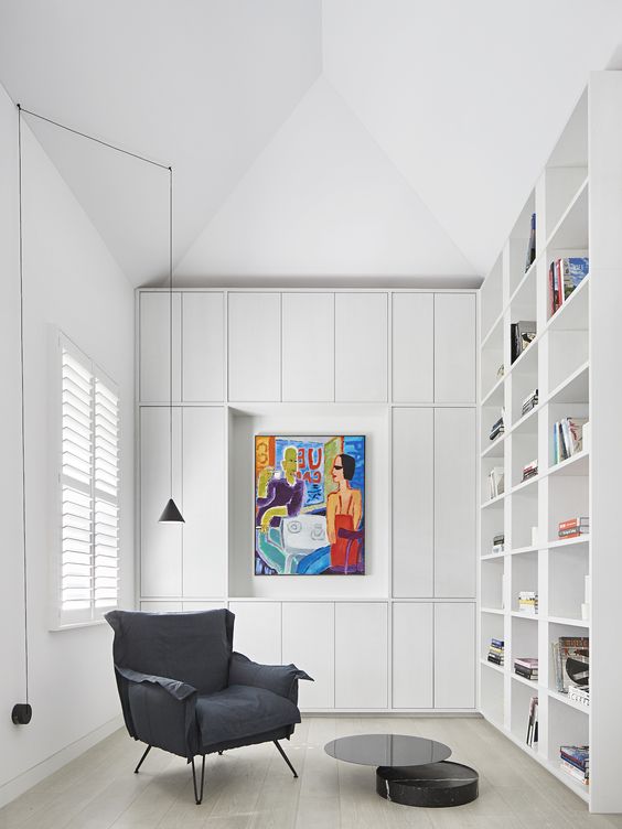 a modern white home library with built-in bookshelves, a grey chair and some closed up storage units