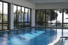 a modern white pool house with all the walls and roof glazed, a large pool and an additional tub plus a stone deck
