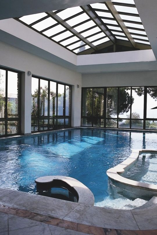 a modern white pool house with all the walls and roof glazed, a large pool and an additional tub plus a stone deck