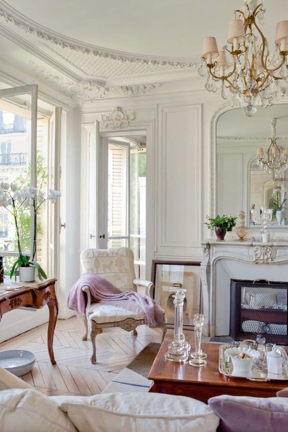 a neutral Parisian living room with a fireplace clad with marble, an antique neutral chair, some antique tables and a crystal chandelier