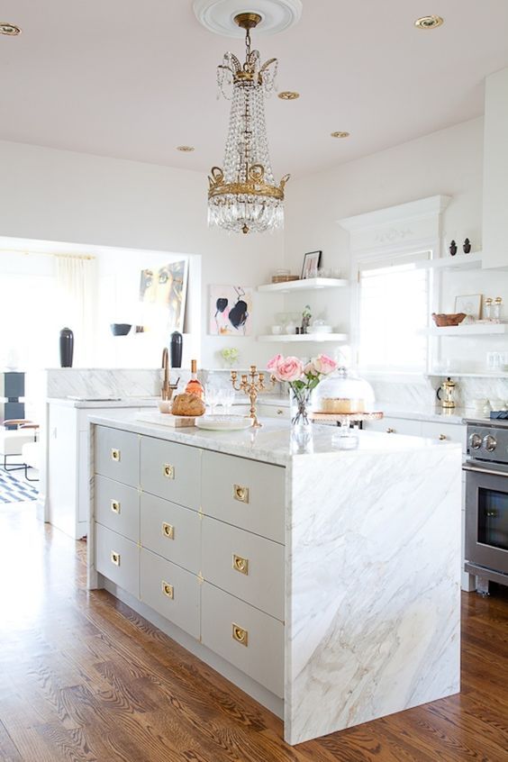 a neutral glam kitchen with white cabinets, white marble countertops and a backsplash, gold handles, a crystal chandelier