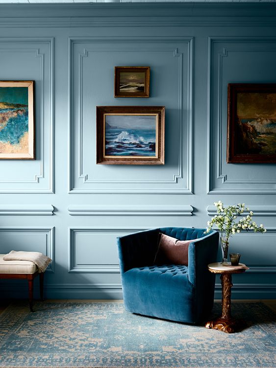 a pastel blue room with molding on the walls, a blue upholstered chair, a gallery wall, some coffee tables and a bench is amazing