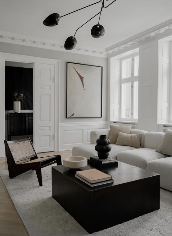 a refined minimalist living room with molding on the walls and doors, a white sectional, a black coffee table, a woven chair, a black chandelier