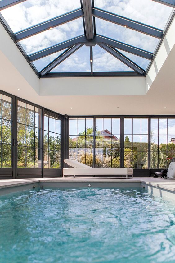 a refined modern pool house with glazed walls and a roog, with lights and a large pool, with some white loungers is amazing