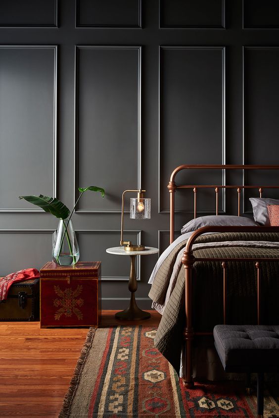 a refined moody bedroom with grey walls with molding, a copper bed, a black bench, a printed rug and a red chest