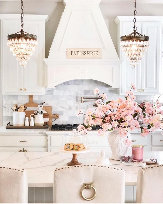 a romantic glam kitchen with white vintage cabinetry, white marble tiles, stools with gold rings, crystal chandeleirs and pink blooms