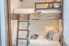 a rustic kids’ bedroom with built-in rough wood beds, a ladder covered with greenery and blooms, built-in sconces and beams on the ceiling