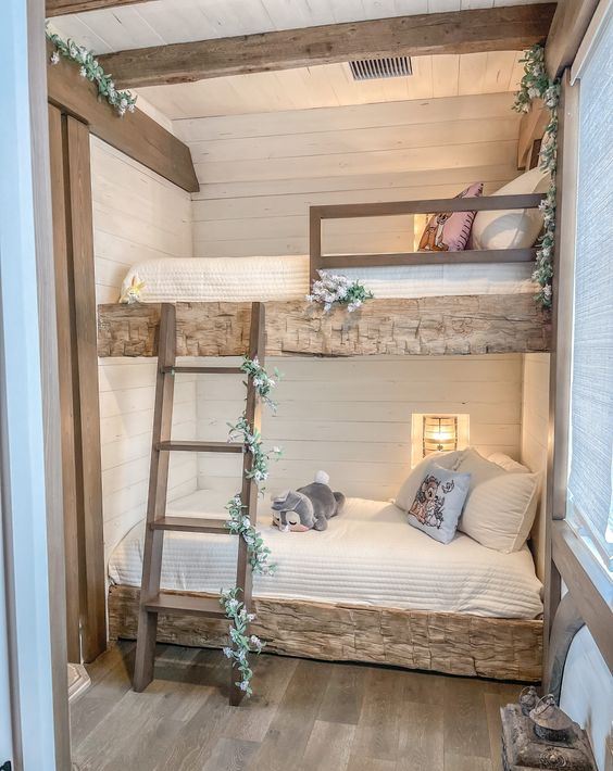 a rustic kids' bedroom with built-in rough wood beds, a ladder covered with greenery and blooms, built-in sconces and beams on the ceiling