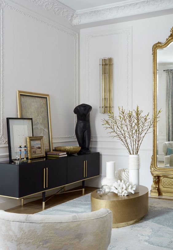 a sophisticated Parisian living room with molding on the walls, gold touches, a black credenza, a gold coffee table and an oversized mirror