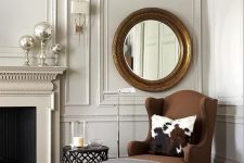 a sophisticated living room with molding on the ceiling and walls, a brown chair, a round mirror in a gilded frame and a non-working fireplace