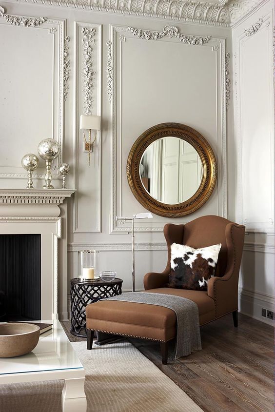 a sophisticated living room with molding on the ceiling and walls, a brown chair, a round mirror in a gilded frame and a non-working fireplace