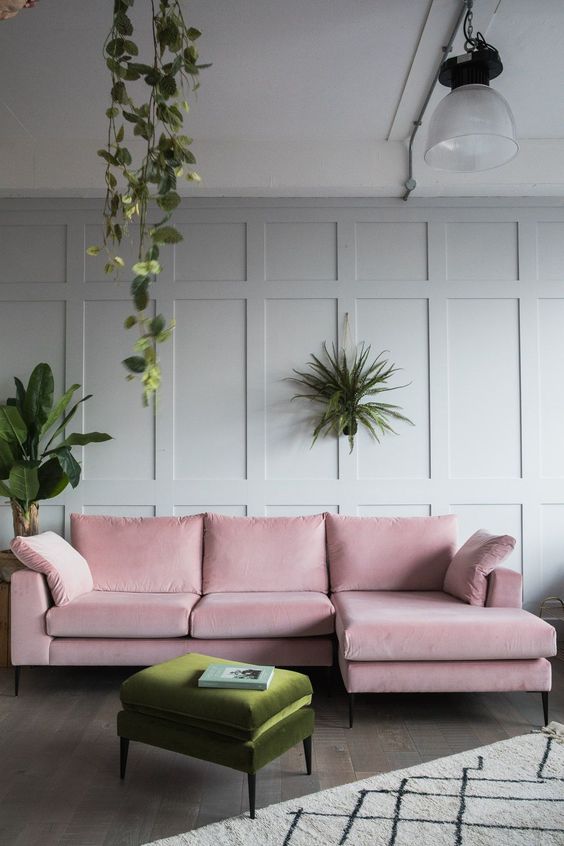 a stylish living room with dove grey paneled walls, a pink sectional sofa, a green pouf and some potted greenery around