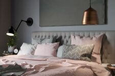 a stylish modern bedroom with grey walls, a grey upholstered bed, grey and pink bedding, a pendant metal lamp and a black sconce