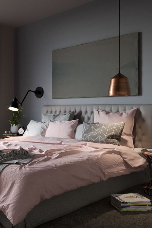 a stylish modern bedroom with grey walls, a grey upholstered bed, grey and pink bedding, a pendant metal lamp and a black sconce