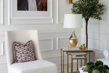 a stylish space with molding on the walls, a creamy chair, a stone coffee table, mini side tables, greenery and touches of gold