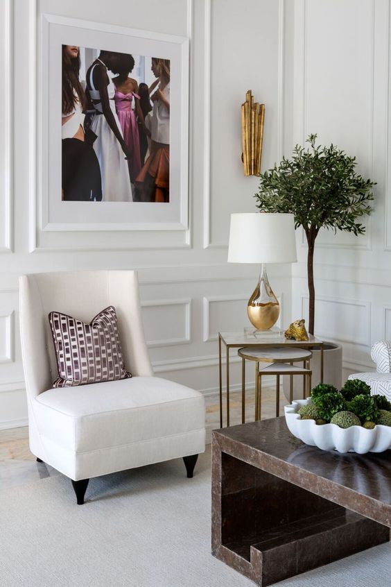 a stylish space with molding on the walls, a creamy chair, a stone coffee table, mini side tables, greenery and touches of gold
