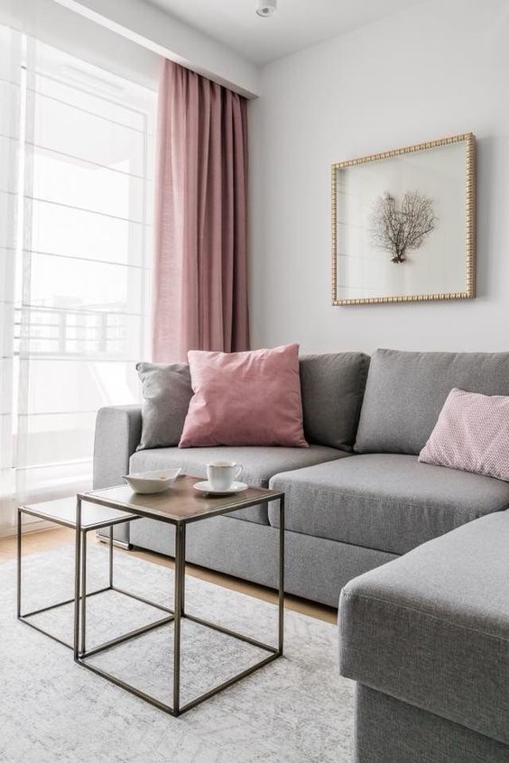 a very laconic living room with a grey sectional, pink and grey sofas, a couple of coffee tables and pink curtains