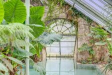 a vintage glass orangery turned into a pool house, with a small pool, lots of greenery, a glass roof and windows