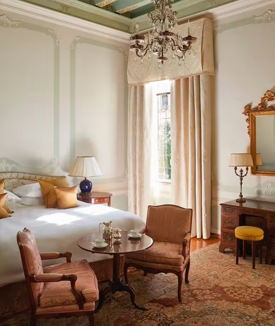 an elegant and refined bedroom with a bed with neutral bedding, a couple of antique pink chairs, a dark-stained desk and a crystal chandelier