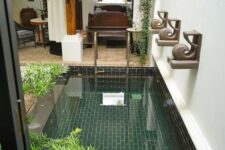 an indoor pool clad with green and black tiles, with potted greenery and some greenery hanging over it is a lovely addition to the space