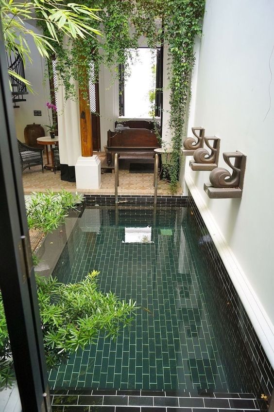 an indoor pool clad with green and black tiles, with potted greenery and some greenery hanging over it is a lovely addition to the space