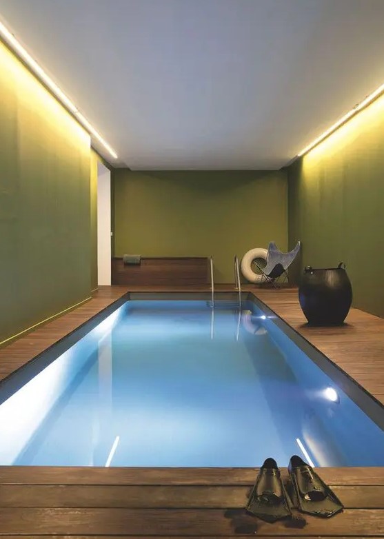 an indoor pool space with green walls, built-in lights, a pool and a wooden deck, a chair and a large vase is a bold idea for a modern home