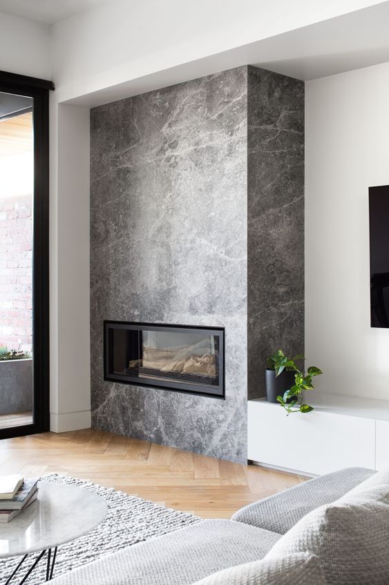 a Scandinavian living room with a built-in fireplace in grey stone, a grey sofa and a rug, a wihte TV unit and a marble coffee table
