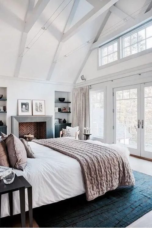 a beautiful and airy bedroom with a brick clad fireplace, a bed with white and dusty pink bedding, niche shelves, wooden beams
